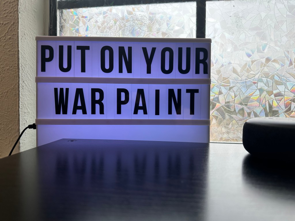 A light up marquee with the words Put on your war paint in large black letters sits one a windowsill. The window behind it is covered with a stain glass privacy film.