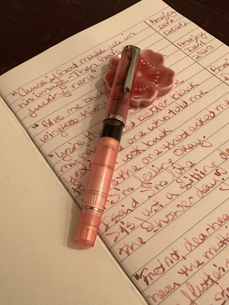 a capped Pelikan m205 rests on a flower pen rest. Underneath them both is an A5 Claire Fontaine notebook. The page is filled with writing in the ink color Rose Quartz. 