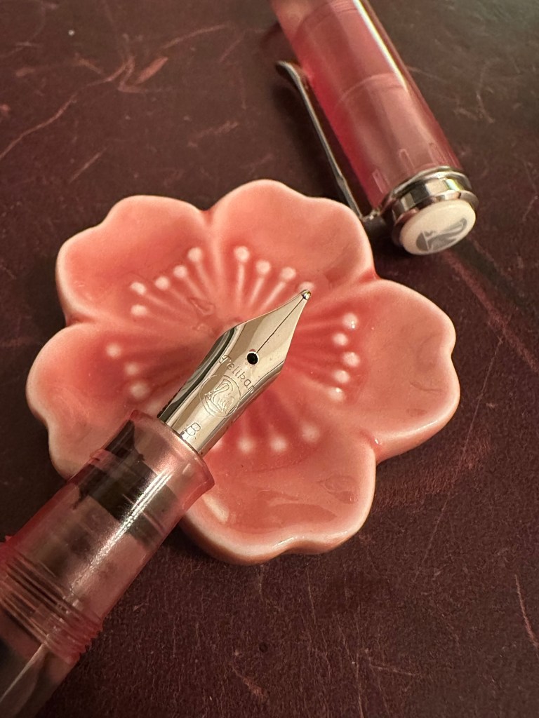 A Pelikan M205 in the color Rose Quartz with a steel broad nib rests on  a ceramic flower pen rest that is also shaded pink.   Just above on the  left hand side you can also see the cap of the pen that has a white finial and silver banding.