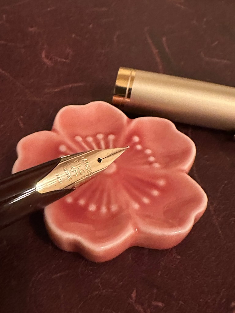 A close up of an inlaid 14 c gold nib on a Pilot E95S.  behind it is a flower pen rest and the cap for the pen.
