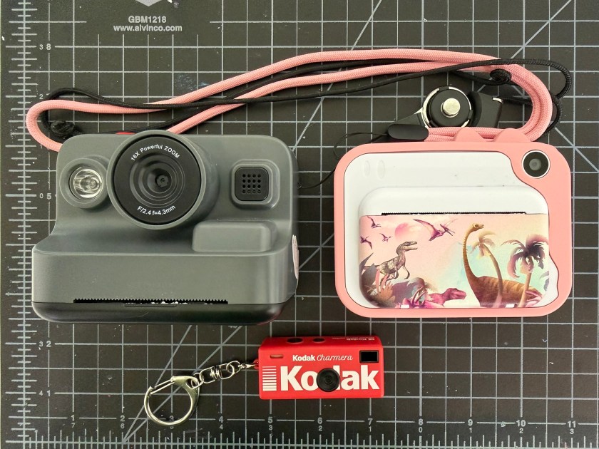 three cameras on black background. the left top one looks similar to a Polaroid and the right one has a scene of dinosaurs wandering across it. Beneath them is a little red Kodak Charmera